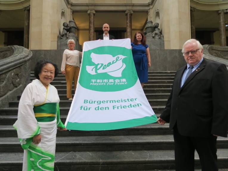 Oberbürgermeister Belit Onay steht mit einem Mann und drei Frauen auf einer Treppe innerhalb des Neuen Rathauses in Hannover. Sie halten die Mayors-for-Peace-Flagge in den Händen.